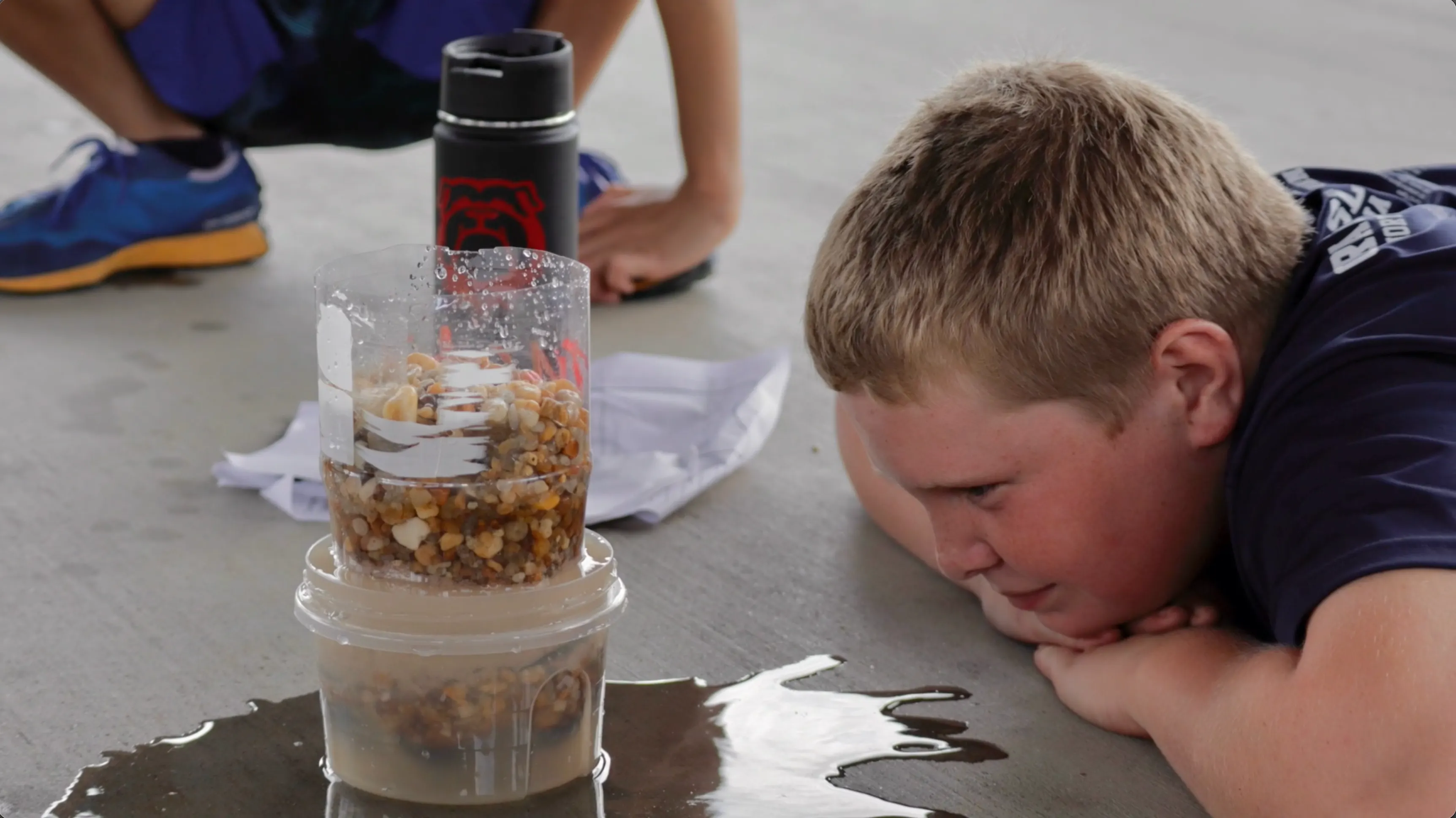 A boy lying flat on the concrete with his chin on his hands, face level with a homemade bottle filter, watching cloudy water drip through into a tub.