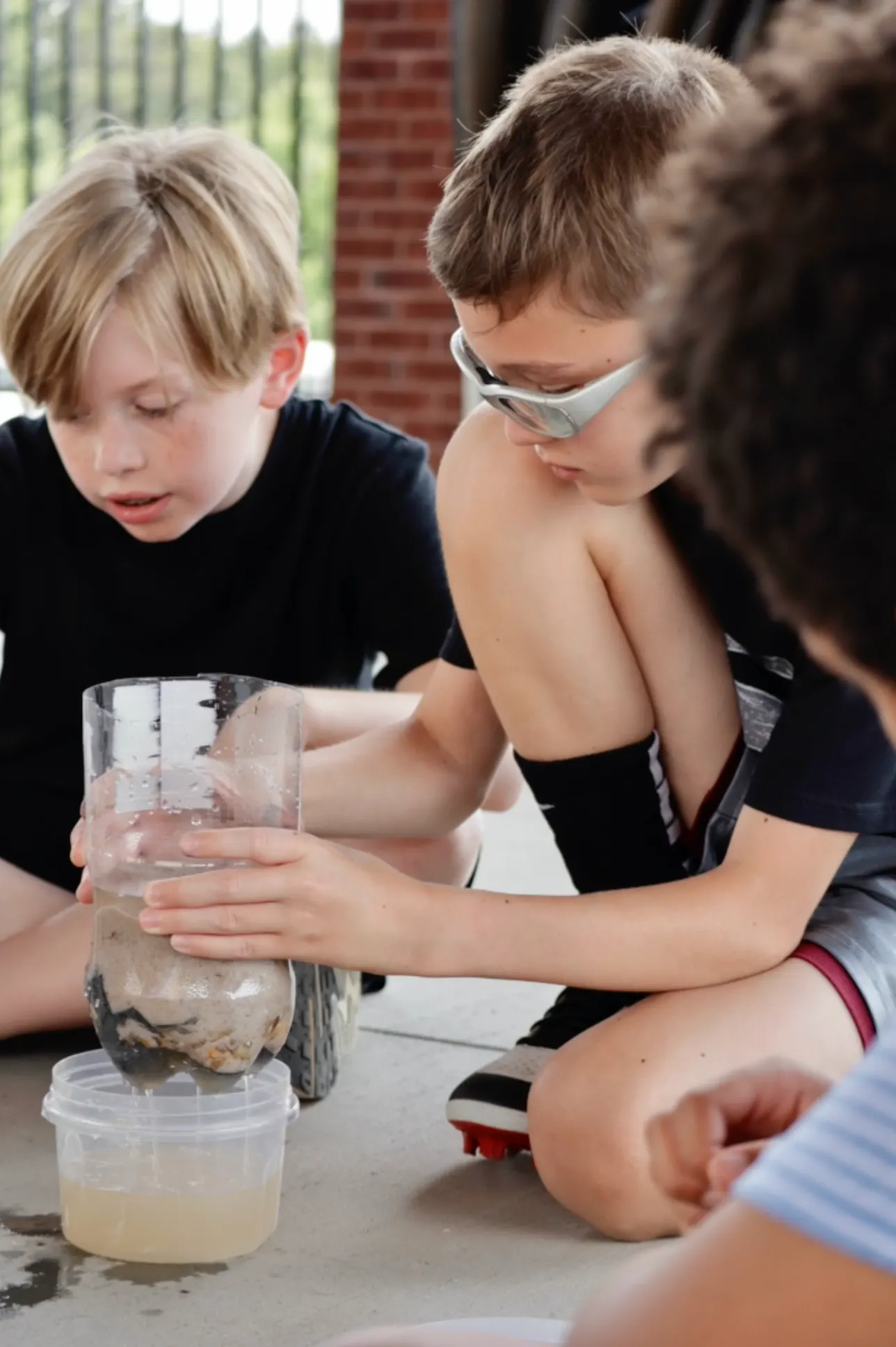 Three children crouched on the ground around a water filter they built from a cut plastic bottle packed with sand and gravel, one holding it steady while it drips into a tub.