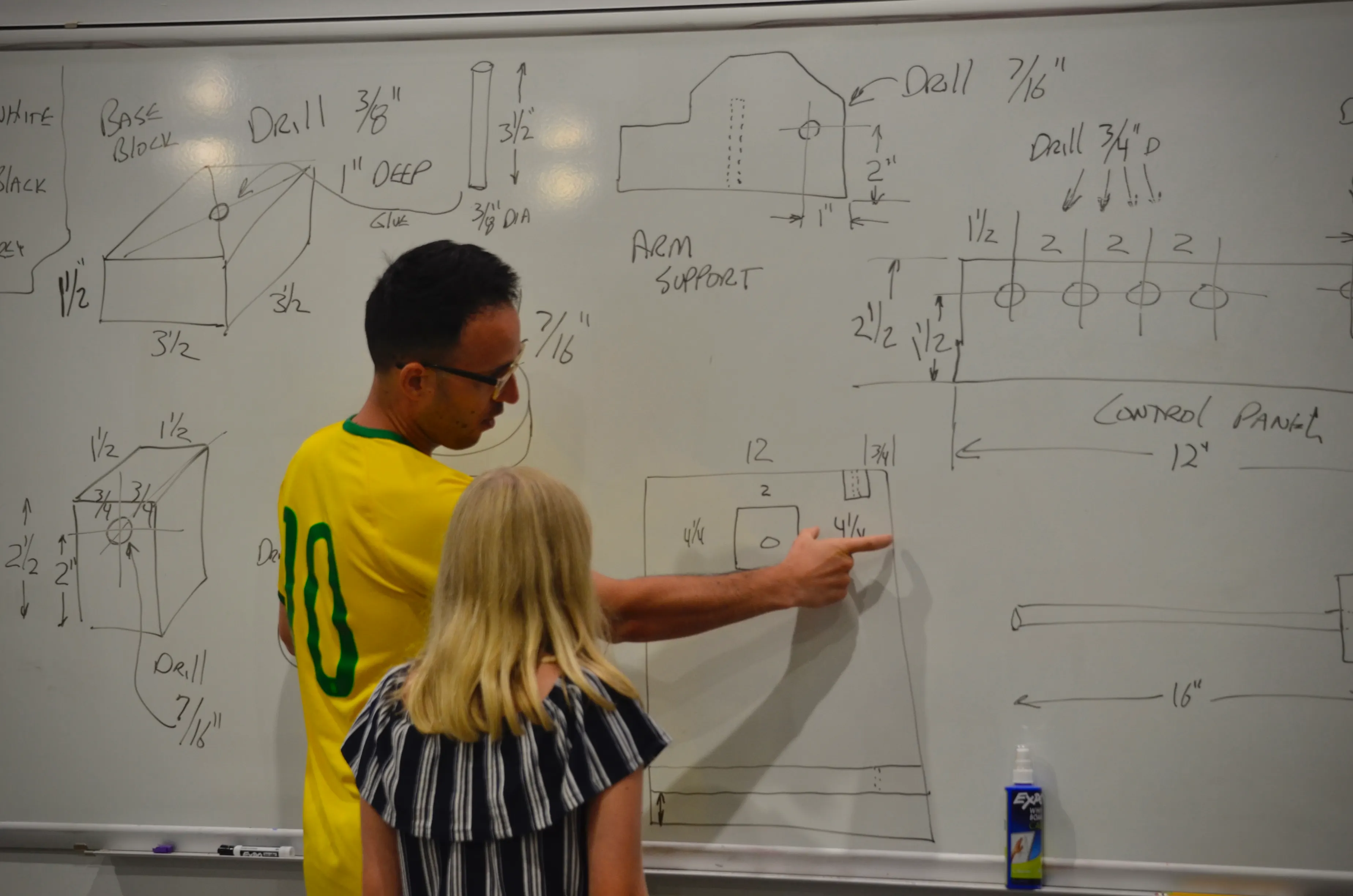 Danny Delgado at a whiteboard covered in dimensioned engineering drawings labelled base block, arm support and control panel, pointing at one detail while a girl stands beside him looking at it.