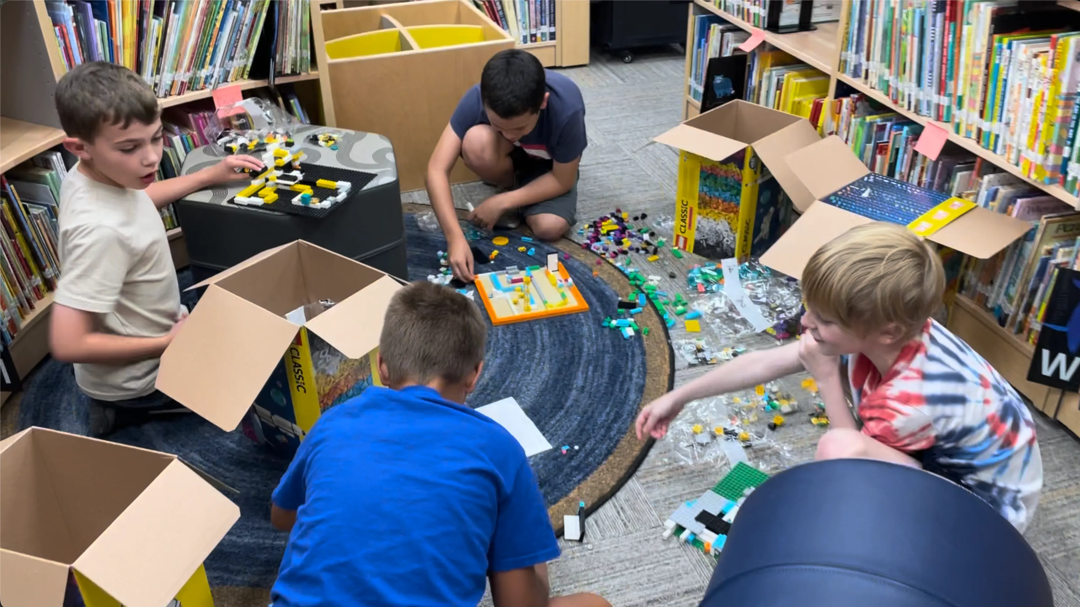 Four boys sitting and kneeling on a library floor between the shelves, building with LEGO spread across a rug, open cardboard boxes and loose bricks all around them.