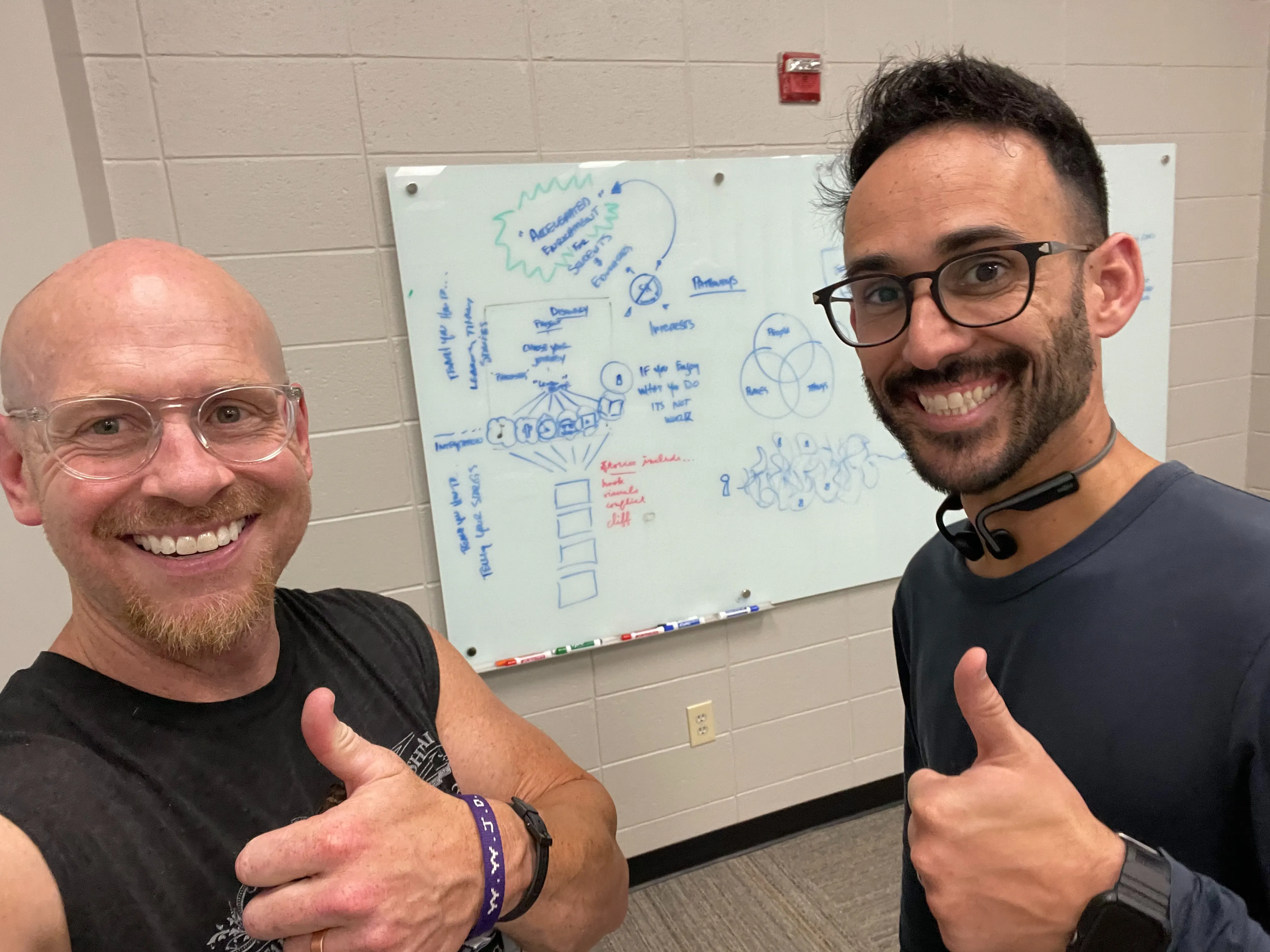 Kevin Wallace and Danny Delgado standing together in front of a glass whiteboard covered in hand-drawn diagrams, both smiling and giving a thumbs up.