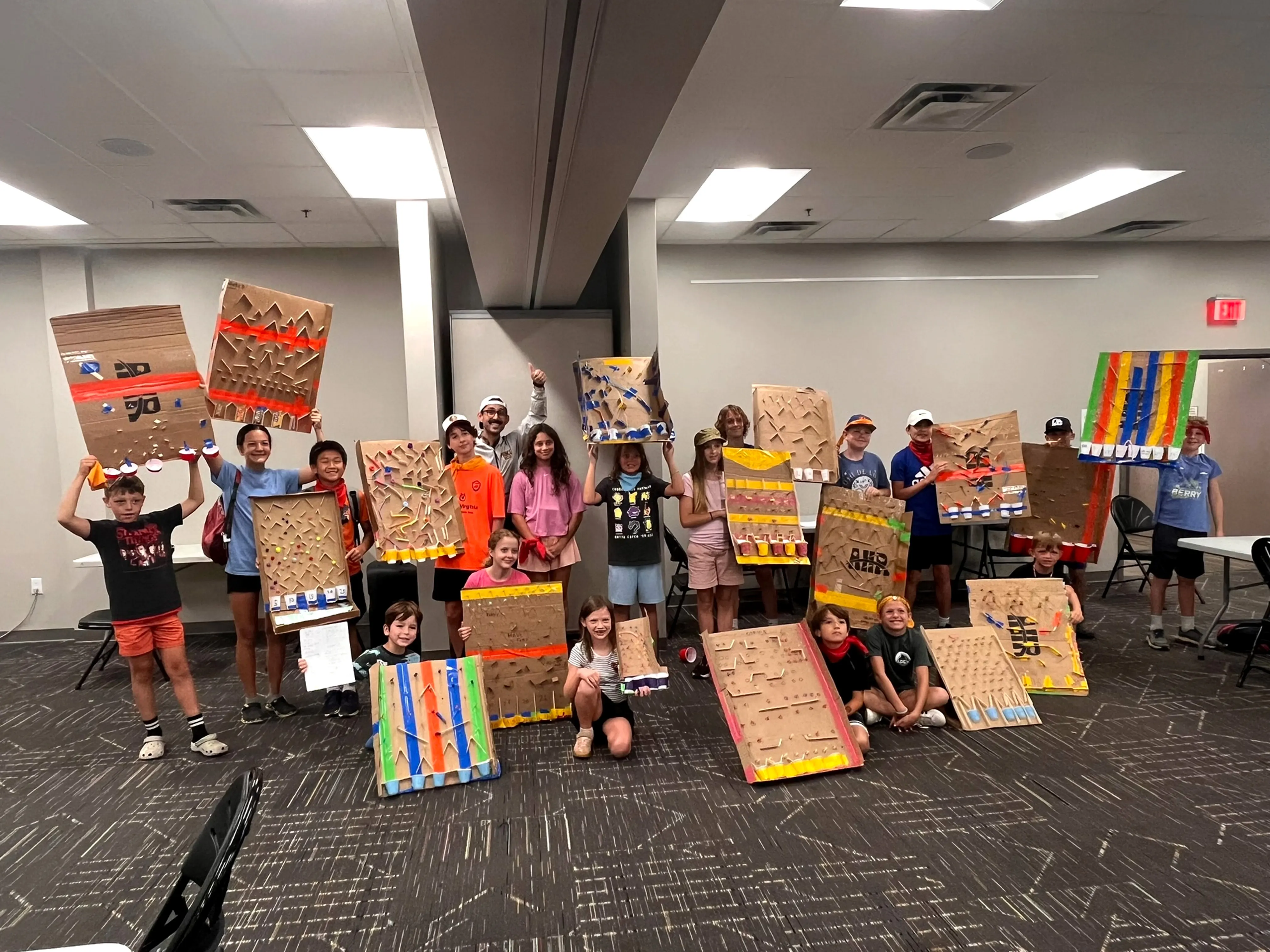 A large group of children holding up cardboard plinko boards they built, each one pegged, painted, and different from the others, with Danny standing among them.
