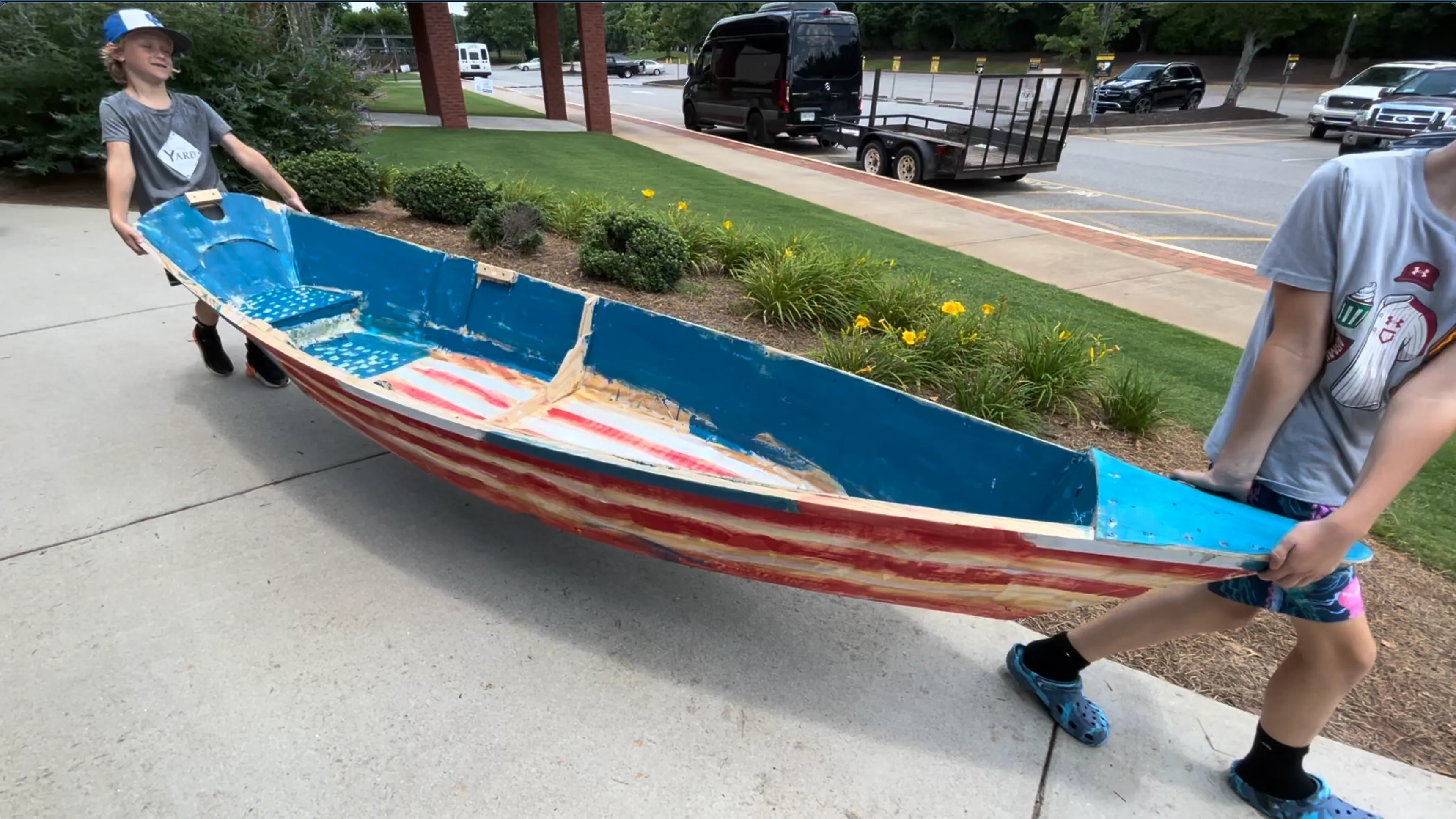 Two boys carrying a full-size wooden boat they built, painted blue on the inside with red and white stripes along the hull, across a sidewalk towards a waiting trailer.